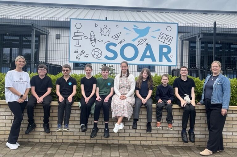 Duke of Lancaster - pupils and teachers sit outside a sign saying 'SOAR'