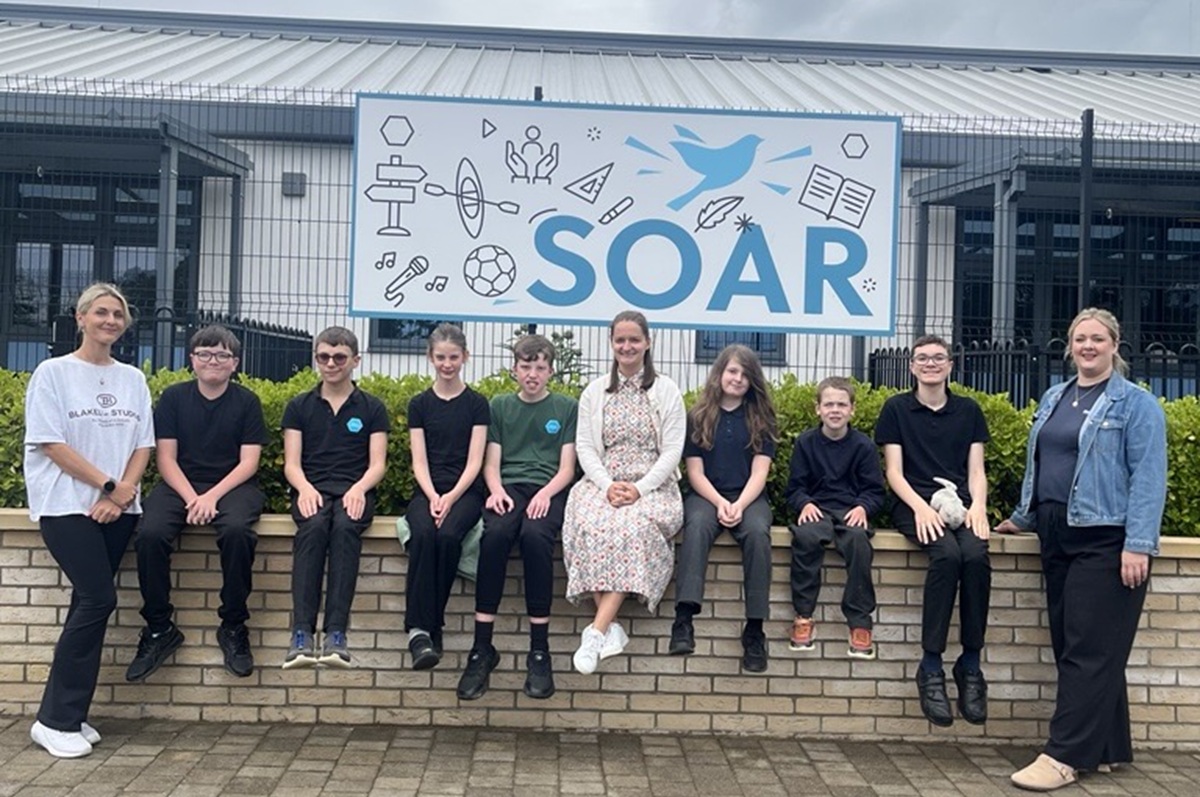 Duke of Lancaster - pupils and teachers sit outside a sign saying 'SOAR'