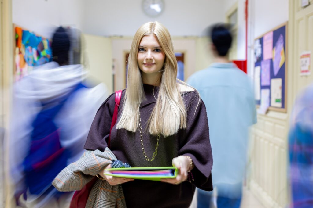Young woman student smiling, holding a binder, standing confidently in a motion blurred school corridor with classmates walking by